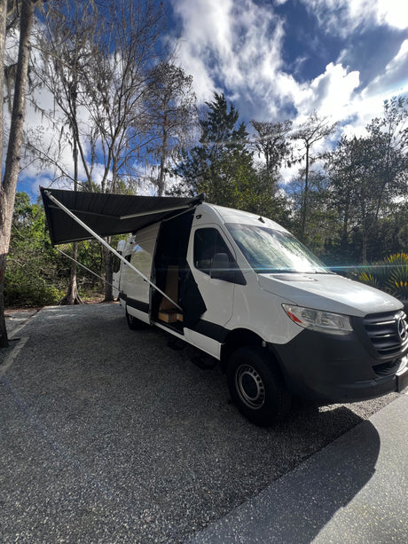 White Sprinter RV rental with extended awning, parked on gravel amidst trees under a bright, cloudy sky.