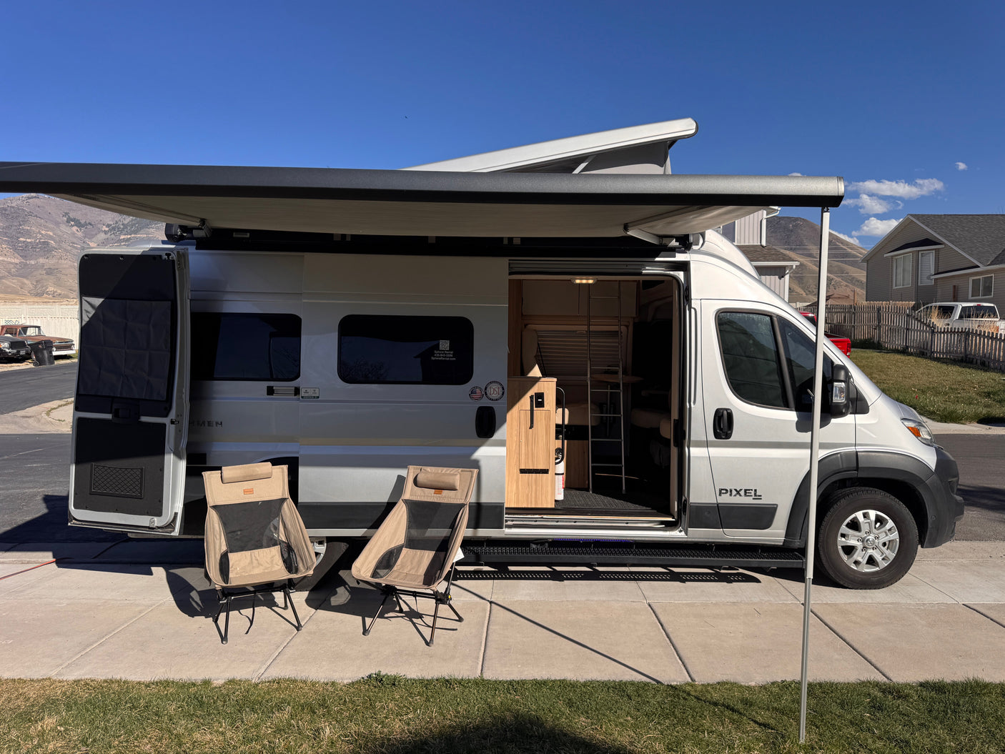 Silver camper van RV rental with an extended awning, open side door revealing the interior, and two camping chairs outside.
