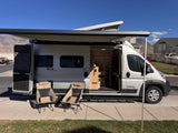 Silver camper van RV rental with an extended awning, open side door revealing the interior, and two camping chairs outside.