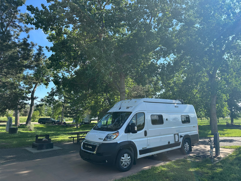 RV rental: A white Winnebago Solis camper van is parked at a shaded campsite, connected to power, with a fire pit nearby. — photo 1