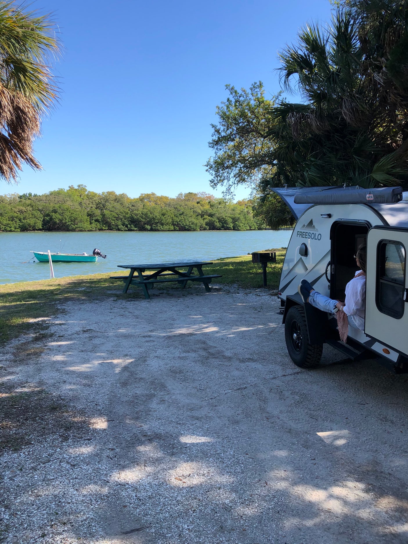 Waterfront RV rental campsite scene: a person relaxing in a FREESOLO camper with a boat & picnic table.