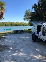 Waterfront RV rental campsite scene: a person relaxing in a FREESOLO camper with a boat & picnic table.
