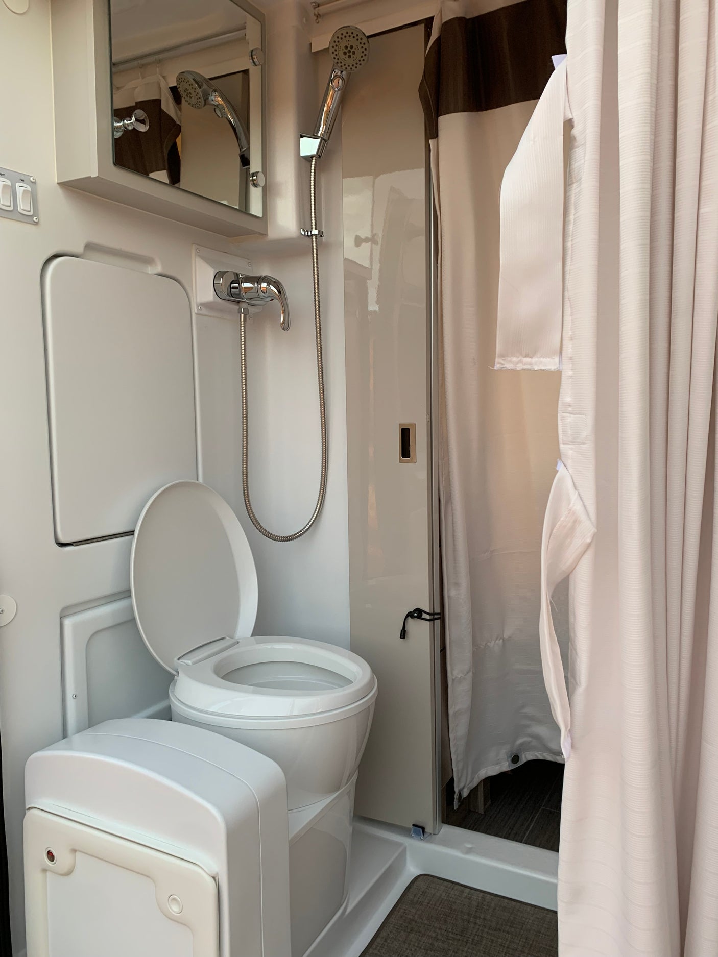 RV rental wet bath: white portable toilet, shower, and mirror. Shower curtain partially open, revealing compact interior.