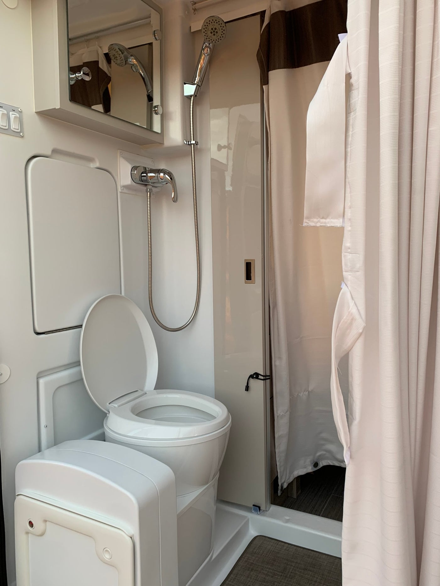 RV rental wet bath: white portable toilet, shower, and mirror. Shower curtain partially open, revealing compact interior. — photo 7
