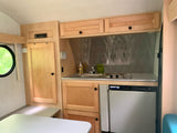 Compact RV rental interior with a functional kitchen featuring a sink, stovetop, refrigerator, light wood cabinets, and dining area.