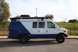 Explore in this custom Ford E-Series camper van, an ideal RV rental. White & blue, with roof storage and off-road tires for adventure.