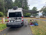 RV rental (Tellaro) parked at a campsite, awning extended, with bikes, scooters, and chairs outside.