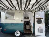This cozy, retro RV rental features a teal and white exterior, striped awning, and a charming interior with a kitchen and bed.