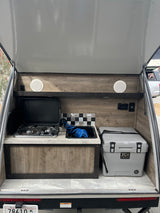 This rv rental features a compact outdoor kitchen with a FlameKing gas stove, sink, and large K2 cooler, ready for camping.
