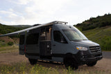 A grey camper van with extended awning and open side door on a dirt path with green hills. Ready for your RV rental adventure.