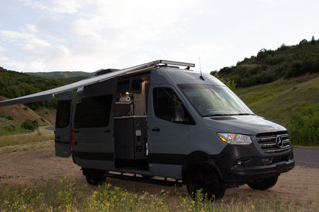 A grey camper van with extended awning and open side door on a dirt path with green hills. Ready for your RV rental adventure.
