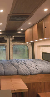 This RV rental interior features a cozy bed, wooden cabinetry, ceiling fan, and a foldable table, with windows offering nature views.