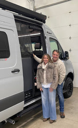 A happy couple poses by their gray camper van's open door, perfect for an upcoming RV rental adventure.