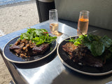 Gourmet beach dining with an RV rental: Two plates of steak & mushroom with salad, and wine, by the sea.