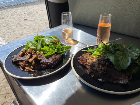 Gourmet beach dining with an RV rental: Two plates of steak & mushroom with salad, and wine, by the sea.