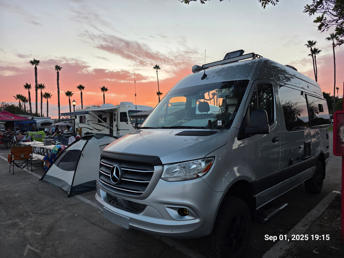 Vibrant sunset over a lively campground, featuring a silver Mercedes Sprinter rv rental among tents and palm trees.