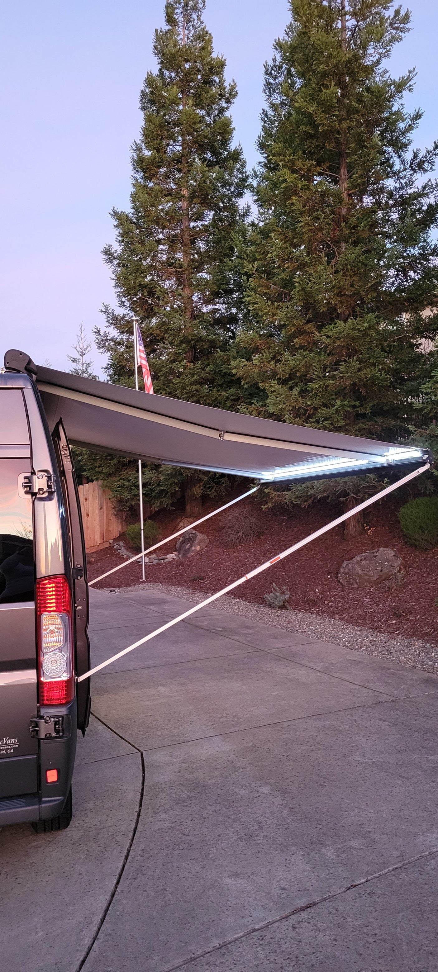 An rv rental camper van with an extended, illuminated awning, supported by poles. Trees, a fence, and a US flag are visible behind.