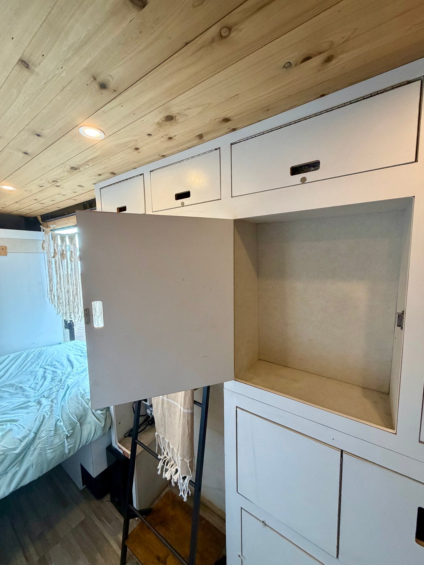 RV rental interior view: Wooden ceiling, bright lights, open white storage cabinet, a bed with blue bedding, and decorative macrame.