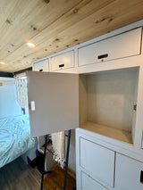 RV rental interior view: Wooden ceiling, bright lights, open white storage cabinet, a bed with blue bedding, and decorative macrame.