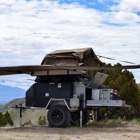 This off-road camping trailer, available for RV rental, offers a rooftop tent, awning, and outdoor kitchen setup.
