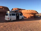 An RV rental, a white pop-top camper van, parked on red desert earth beside massive red rock formations under a clear blue sky.
