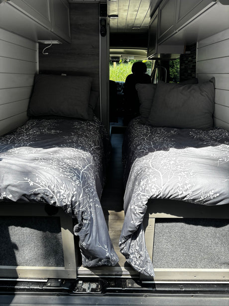 RV rental camper van interior with two comfortable single beds, grey patterned bedding, and narrow aisle leading to the front.