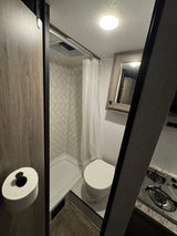 Inside an rv rental: a compact bathroom with shower and toilet, adjacent to a small kitchen area with a stove and mirrored cabinet.
