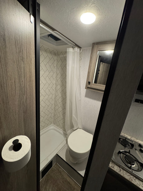 Inside an rv rental: a compact bathroom with shower and toilet, adjacent to a small kitchen area with a stove and mirrored cabinet.