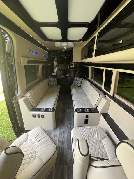 Inside a modern rv rental: cream quilted leather seats, dark wood floor, overhead storage, and accent lighting.