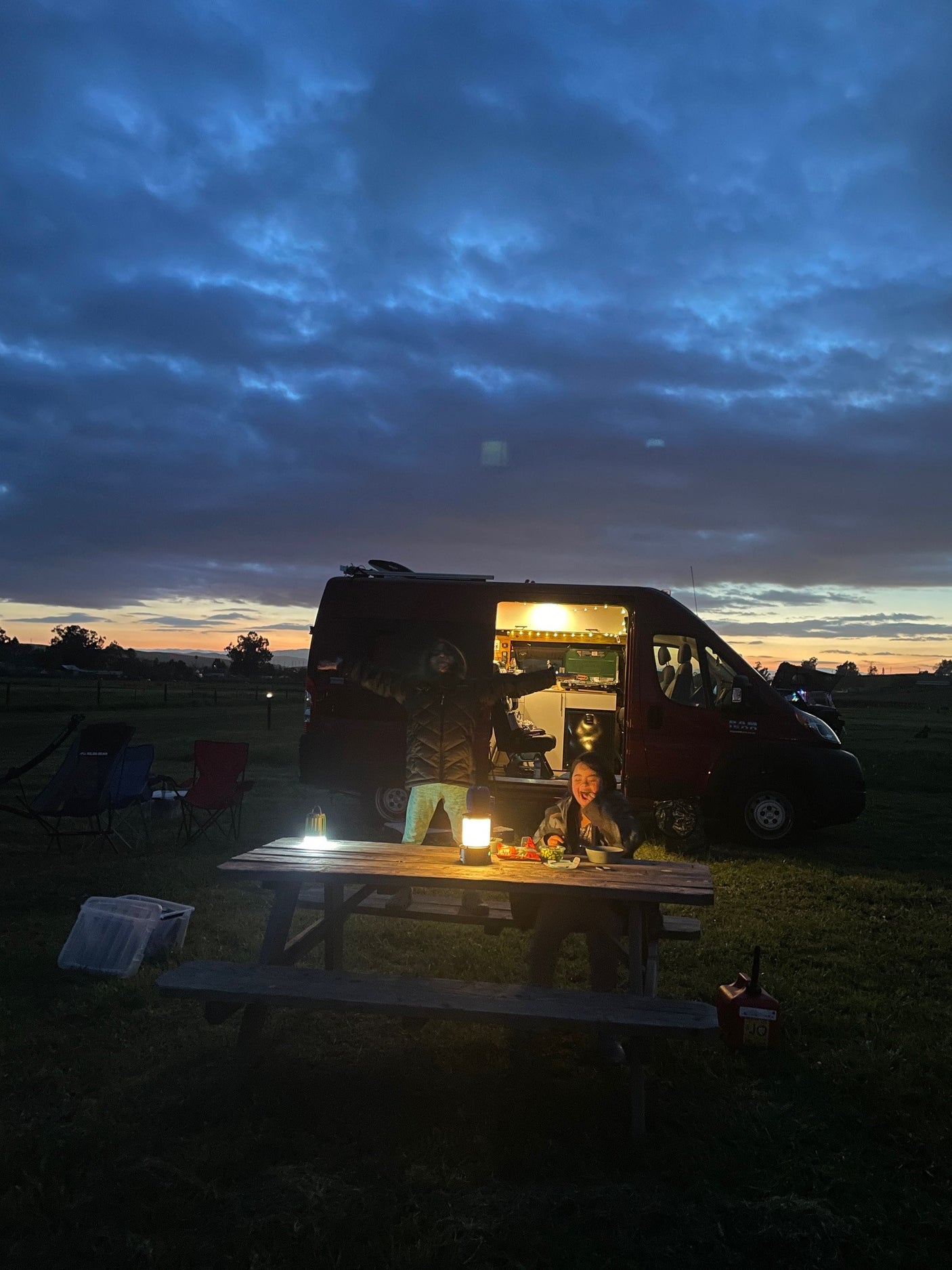Dusk camping with an RV rental: Two people, a red camper van, and a lit picnic table under a colorful sunset sky.