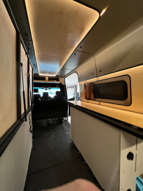 Experience this custom RV rental: a bright camper van interior with warm strip lighting, a functional counter, and spacious aisle.