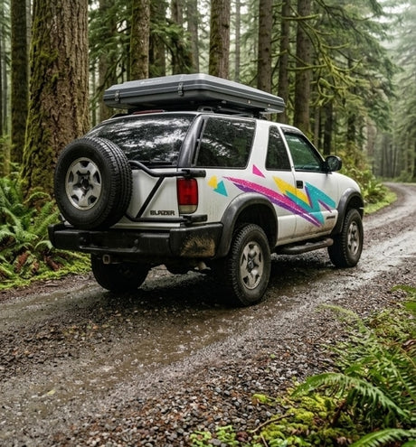 A white Chevy Blazer with a rooftop tent and vibrant decals on a forest trail. Ideal for your next RV rental adventure!