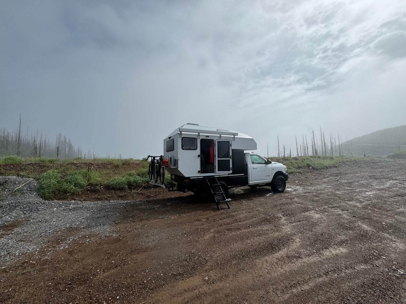 White truck camper RV rental parked on a dirt road amidst foggy, treeless landscape. Perfect for rugged outdoor adventures.