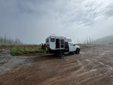 White truck camper RV rental parked on a dirt road amidst foggy, treeless landscape. Perfect for rugged outdoor adventures.