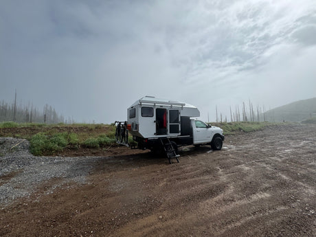 White truck camper RV rental parked on a dirt road amidst foggy, treeless landscape. Perfect for rugged outdoor adventures.