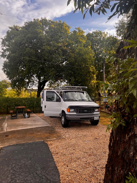 This white camper van, ideal for your next rv rental adventure, is parked on a gravel driveway with its door open next to a picnic table.