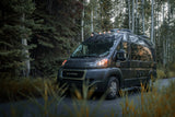 Dark grey RAM ProMaster RV rental van by evergreens & aspens, lights on, parked on a forest road with tall grass.