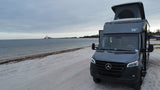 Grey pop-top RV rental on sandy beach by water with bridge view. Perfect for your next adventure!