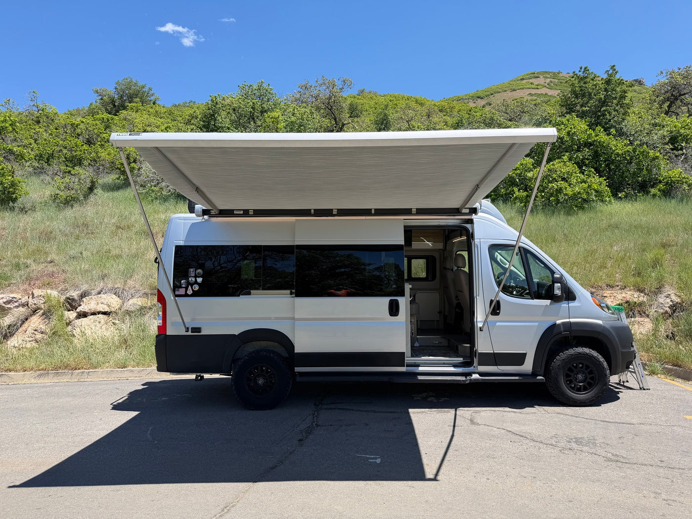 Silver RV rental camper van, awning extended, side door open, showing interior. Parked on asphalt by a green hill on a sunny day. — photo 4