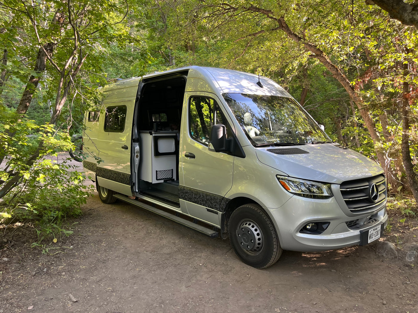 Silver Mercedes-Benz Sprinter rv rental van with its side door open, parked on a dirt path in a lush green forest. — photo 3