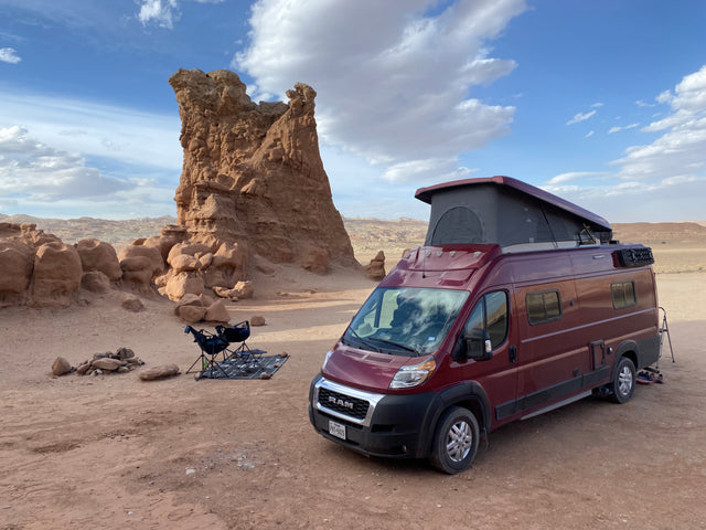 Red RV rental campervan parked among unique desert rock formations, with camping chairs and fire pit under a blue sky.