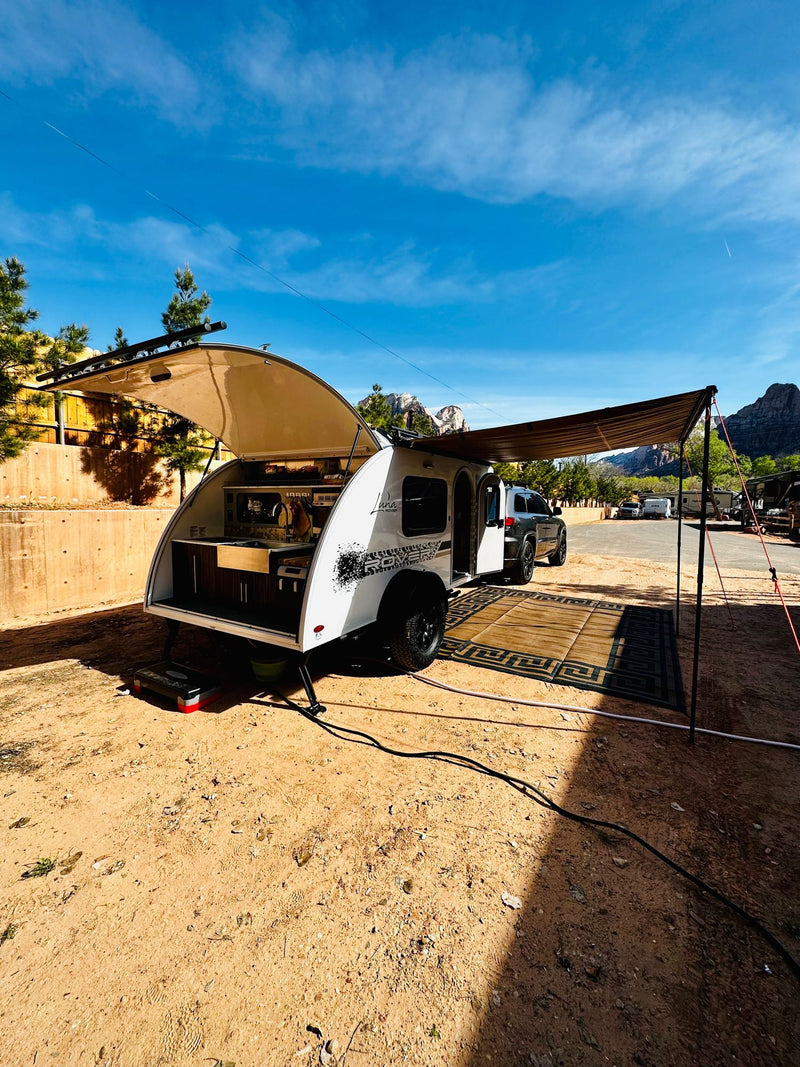  in a parking lot at sunset.|This RV rental is a white teardrop camper with an open outdoor kitchen and awning — photo 9