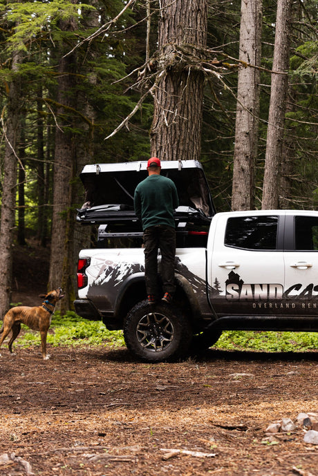 Man opens rooftop tent on an RV rental truck in a forest with a dog. Perfect for overland adventures.
