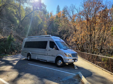 Silver Mercedes-Benz Sprinter rv rental parked on asphalt, surrounded by sunny autumn trees and a metal safety railing.