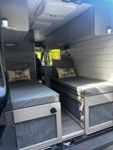 Cozy RV rental interior with twin beds, gray cushions, overhead storage, a compact kitchen, and ceiling AC unit.