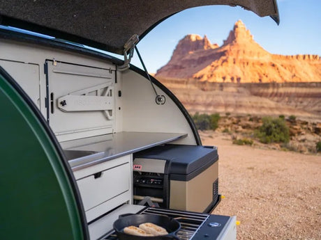 An RV rental teardrop trailer's rear kitchen, with stove, fridge & sink, open to a stunning desert mountain landscape.