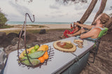 Perfect RV rental beach day: two people relaxing under trees, toasting beers, with a picnic table of fruit and cheese.