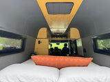 RV rental interior: cozy bed, modern wood ceiling, & side windows offer views. Looking from rear to front seats.