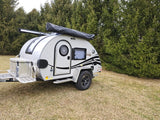 This compact teardrop RV rental, with an awning and storage box, is ready for adventure. Parked on grass with trees in background.