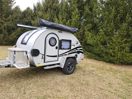 This compact teardrop RV rental, with an awning and storage box, is ready for adventure. Parked on grass with trees in background.